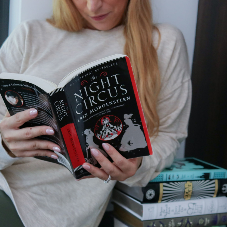 Woman reading The Night Circus with a stack of books beside her, exploring book club discussion questions and resources.