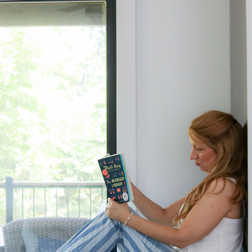 Woman sitting by window with The Midnight Library, enjoying cozy reading time and bookish lifestyle.