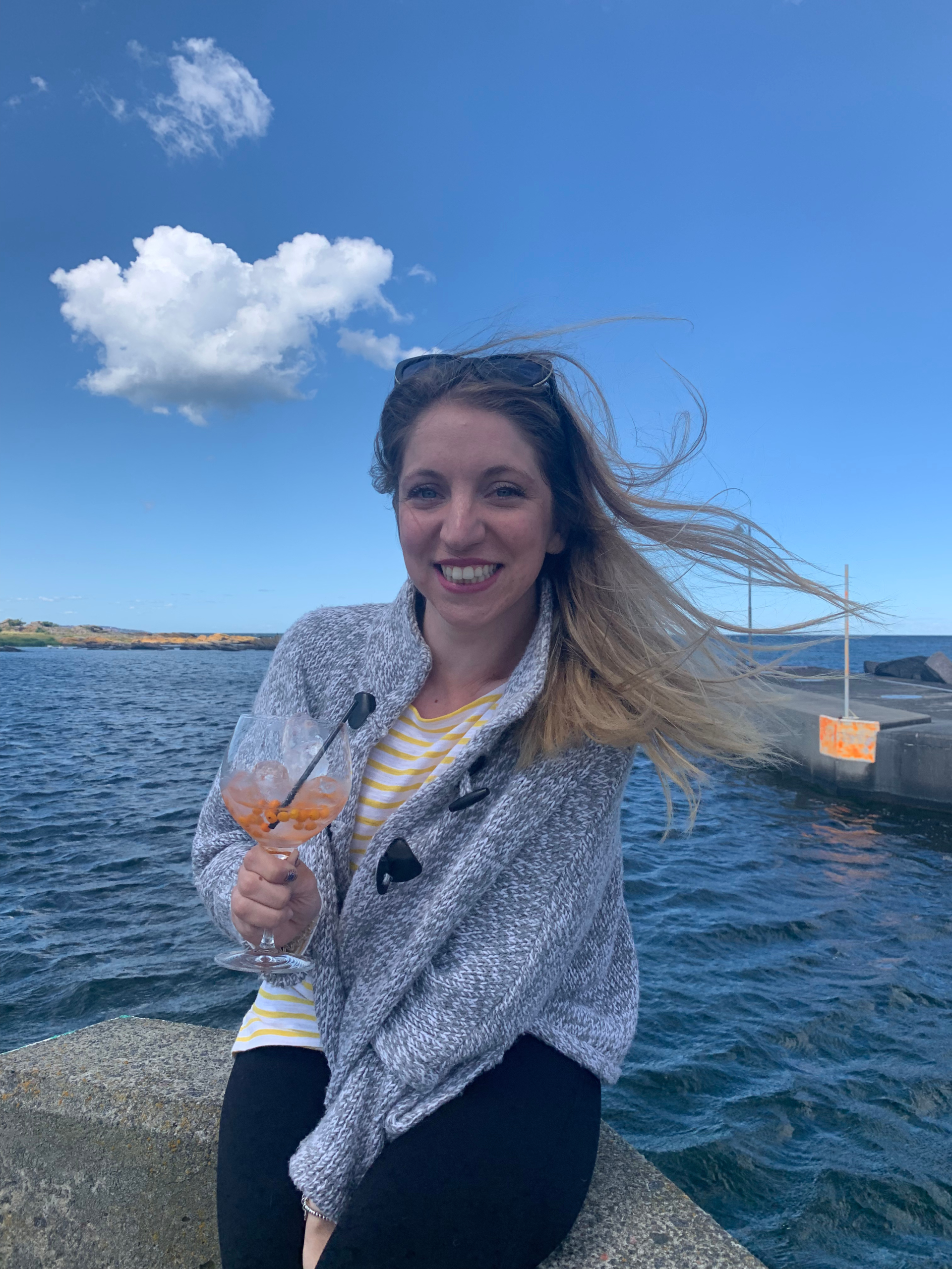 Woman sitting by the ocean in a sweater, holding a cocktail and smiling