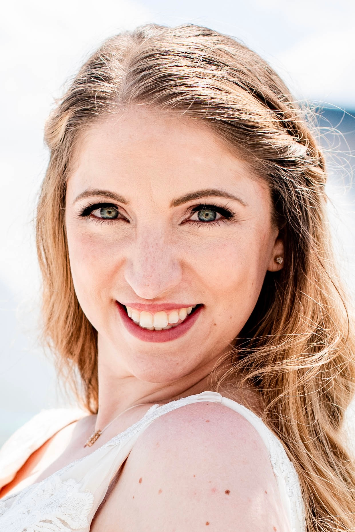 Close-up of smiling woman in bridal makeup and white dress, outdoors with windblown hair