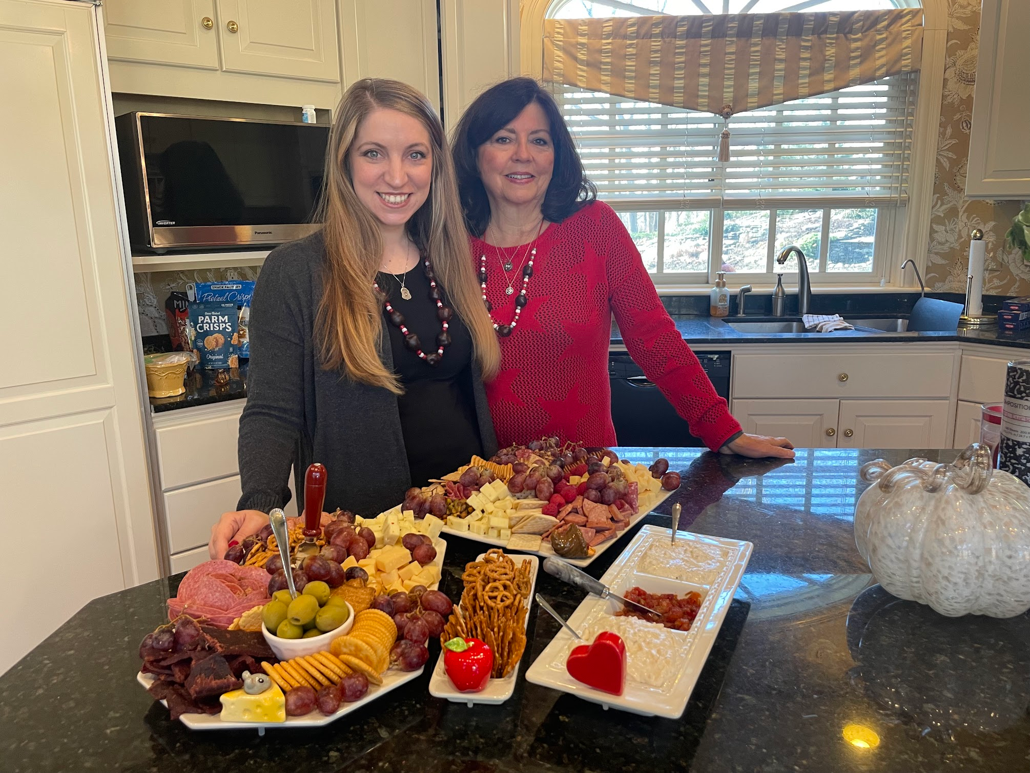 Two women in a kitchen smiling behind a charcuterie spread with cheese, fruit, and crackers — perfect inspiration for book club hosting and cozy book vibes.