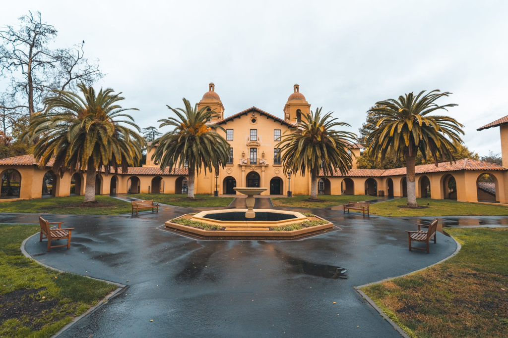 Front view of a Stanford University building with palm trees and fountain, capturing the academic setting in Deep End by Ali Hazelwood, a campus sports romance book.